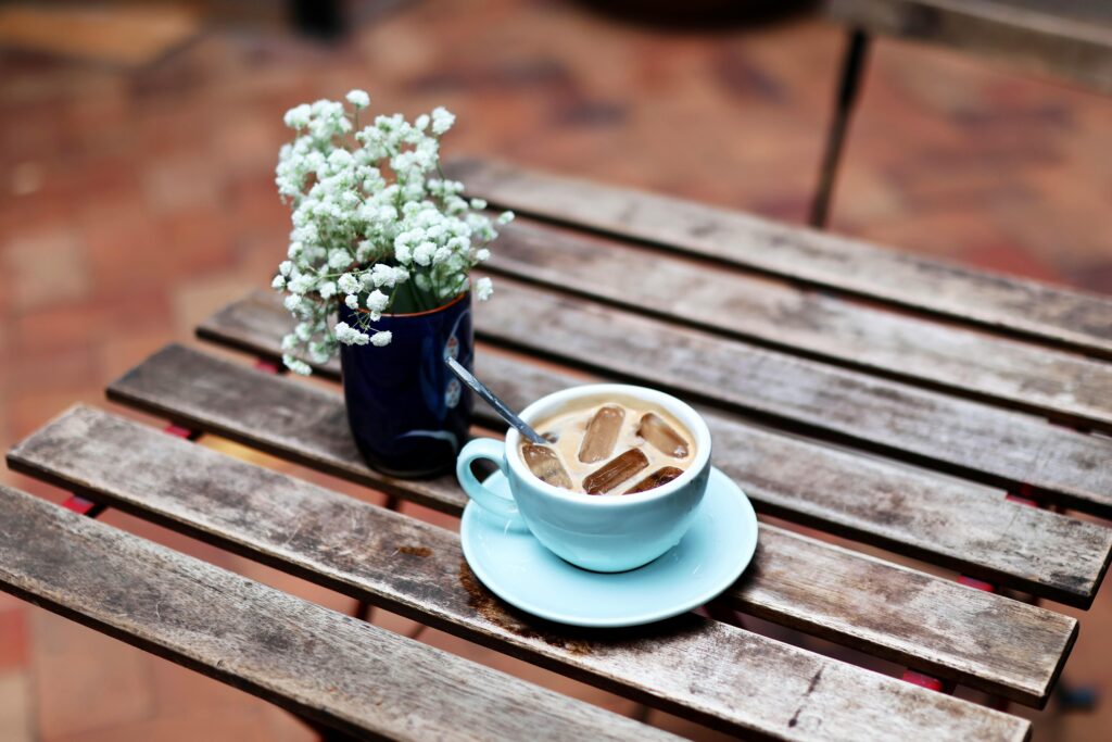 Iced coffee in a blue cup on a rustic table with flowers in a vase outdoors.
