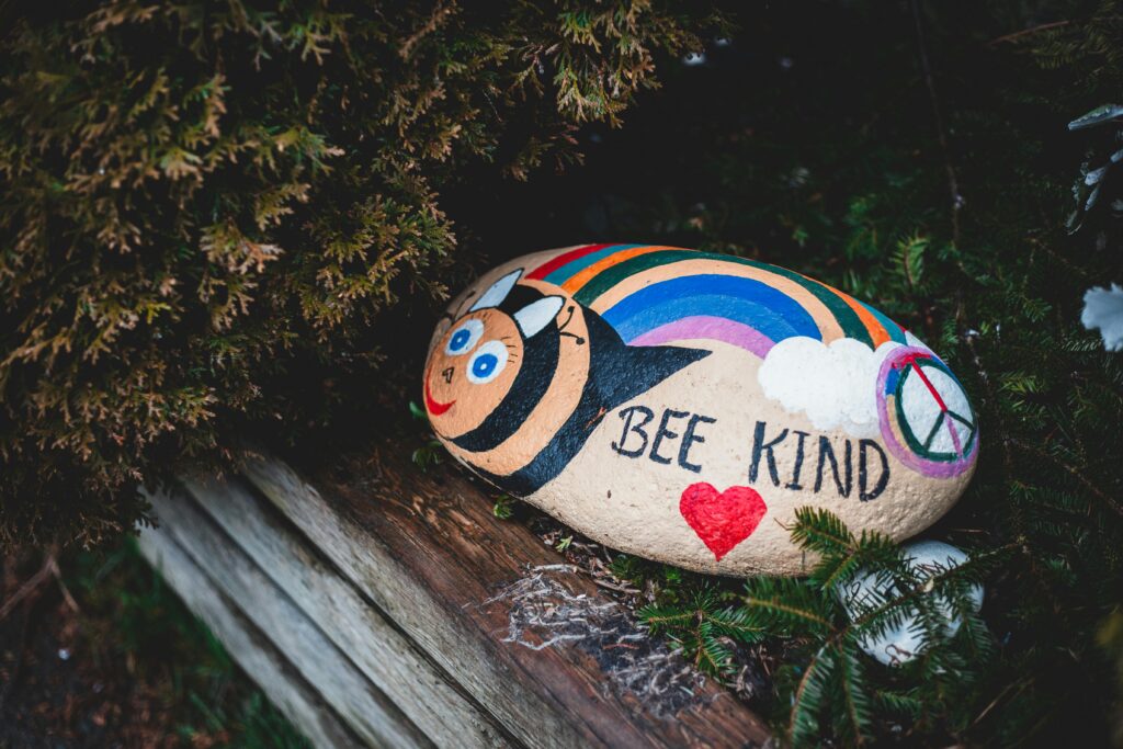 Colorful painted stone with 'Bee Kind' message, bee design, and rainbow in a natural setting.