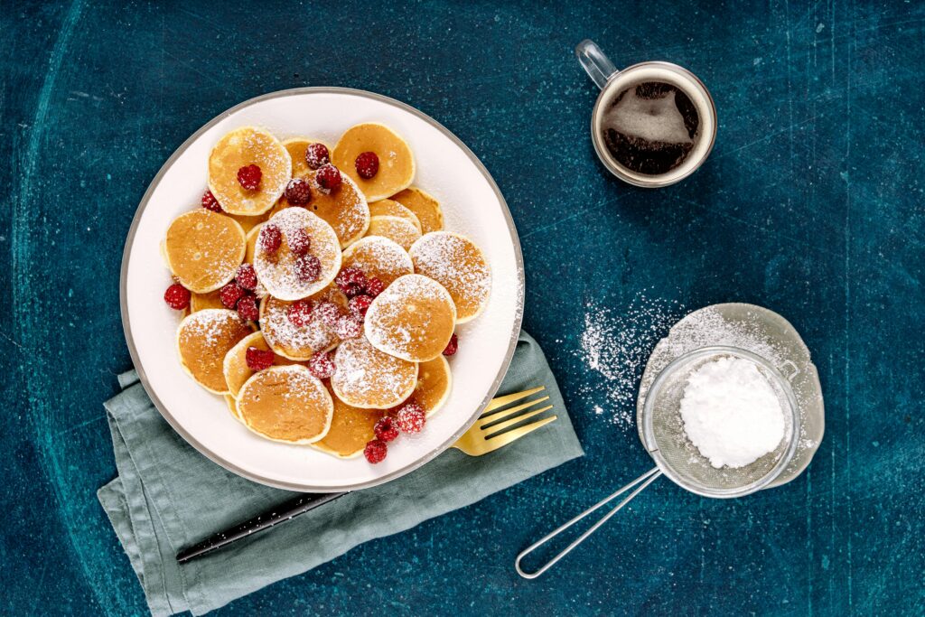 Overhead view of mini pancakes with raspberries, powdered sugar, and coffee on a blue background.