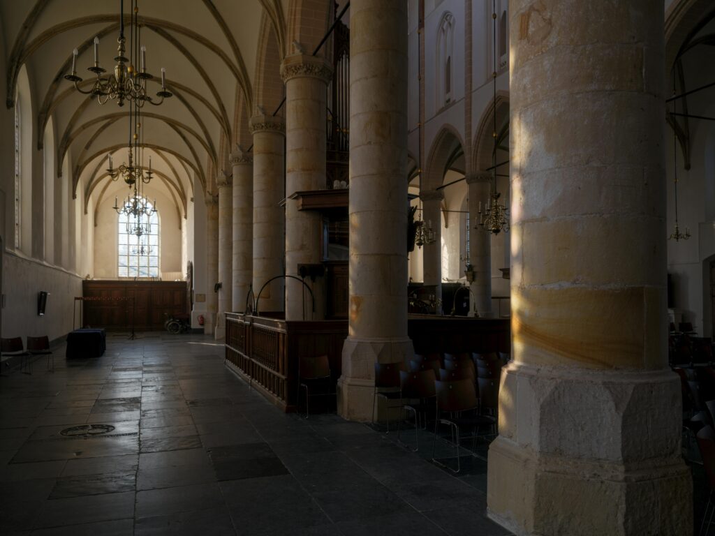 Interior view of a gothic church with stone columns, arches, and sunlight streaming through a large window.