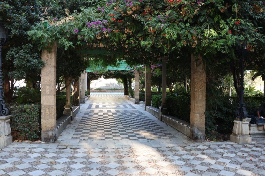 Explore a picturesque garden walkway in Cádiz, Spain, surrounded by lush greenery and vibrant flowers.